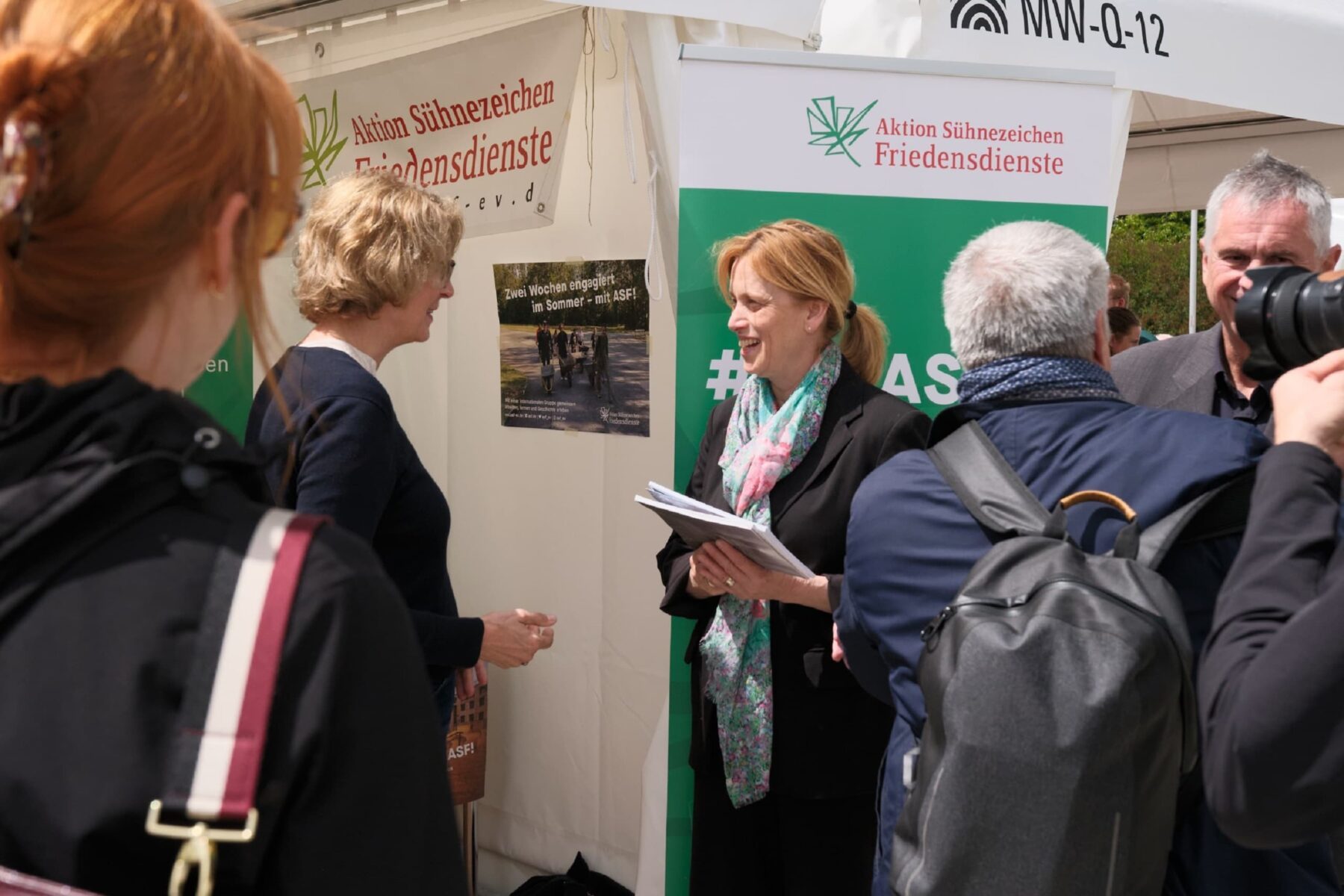 Frau hält Unterlagen am Stand der Aktion Sühnezeichen Friedensdienste vor grünem Banner, im Gespräch mit Besuchern.